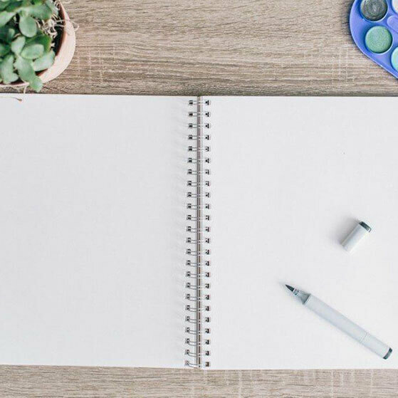 Blank spiral notebook on a wooden desk with a small plant, paint palette and pen.