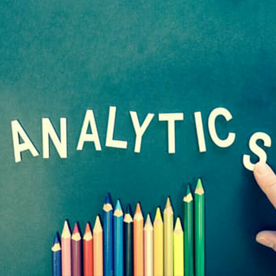 ANALYTICS spelled out above a row of colored pencils, with a finger pointing at the last letter.