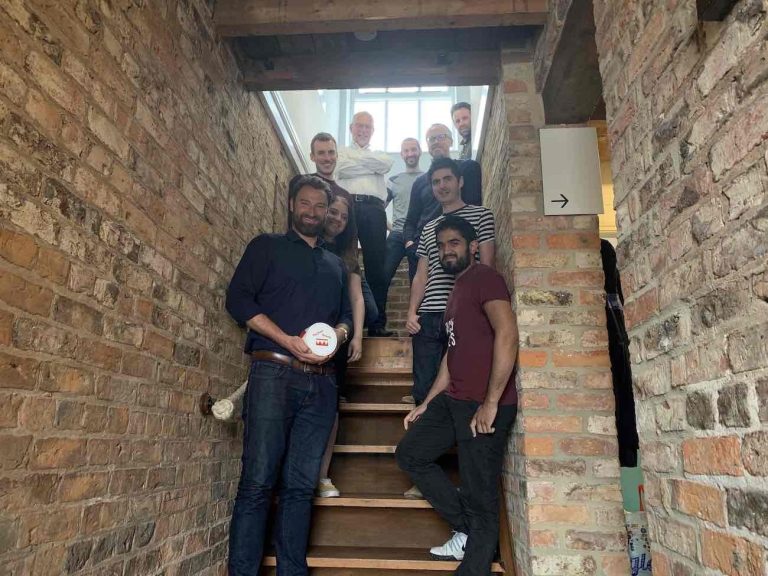 Seven men posing on a wooden staircase in a brick-walled interior.