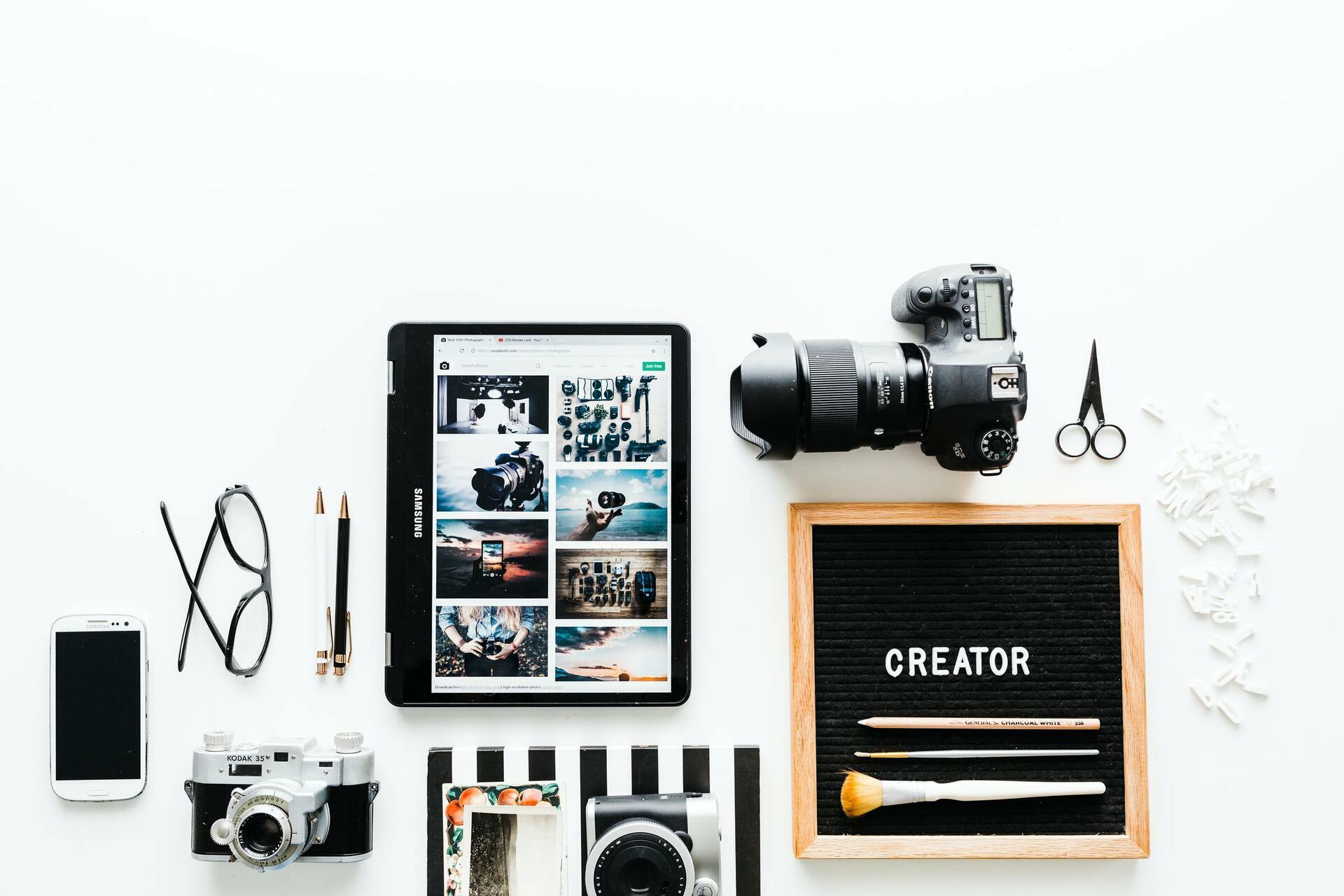 Flat lay of creator tools, including a tablet with a photo grid, camera, phone, glasses, and a board reading “CREATOR”.