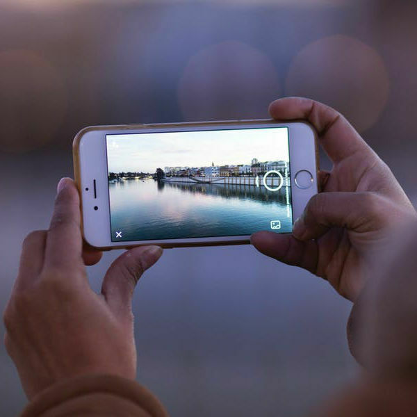 Hands holding a smartphone to photograph a waterfront cityscape at dusk.