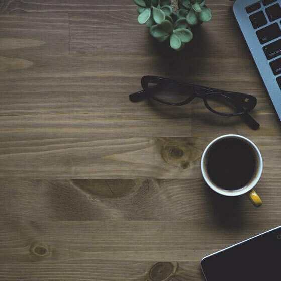 Wooden desk with coffee cup, eyeglasses, laptop, phone and a small plant.