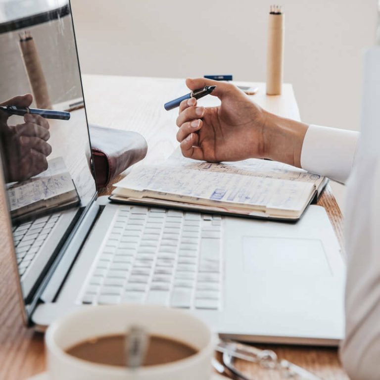 Hand holding a pen over notes beside a laptop and coffee cup.