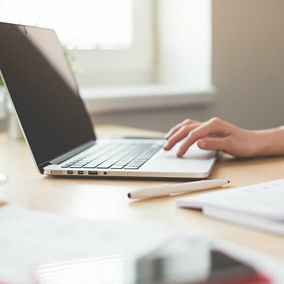 Hand typing on a laptop at a desk beside a pen and papers.