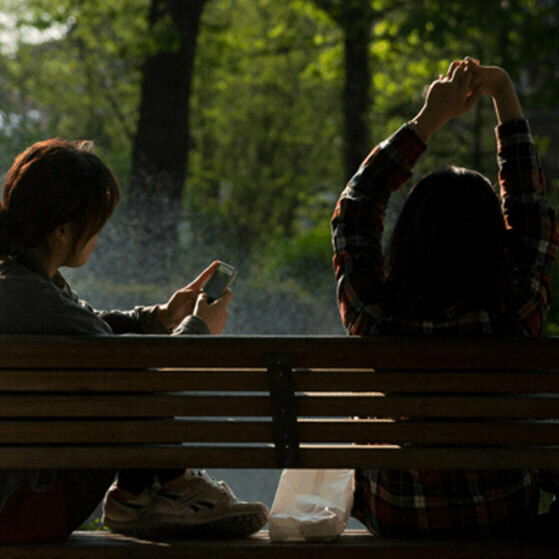 Two people sit on a park bench, one checking a phone and the other stretching overhead.