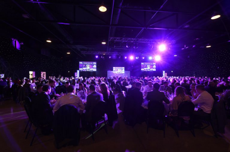 Audience seated at tables in a purple-lit conference hall facing a stage with screens.