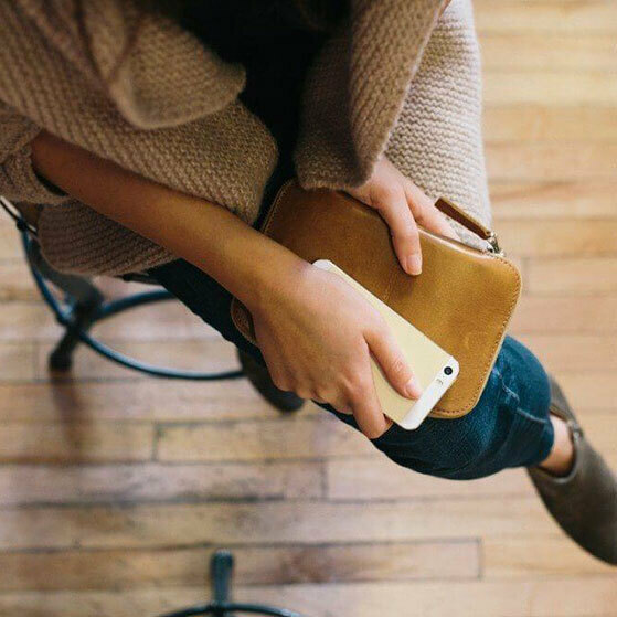 Person sitting on a chair holding a white smartphone and a tan clutch bag.