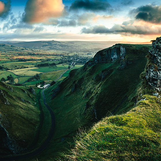 Steep green hillside cliffs overlooking a patchwork valley under a dramatic cloudy sky.
