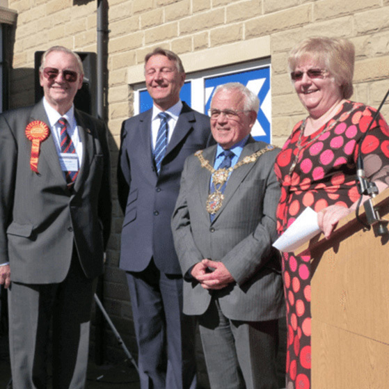 Four officials in suits and a woman at a podium stand outside a building at a ceremonial event.