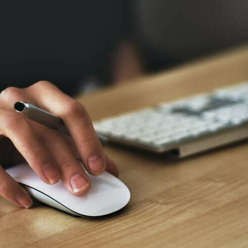 Hand using a white computer mouse beside a keyboard, holding a pen.