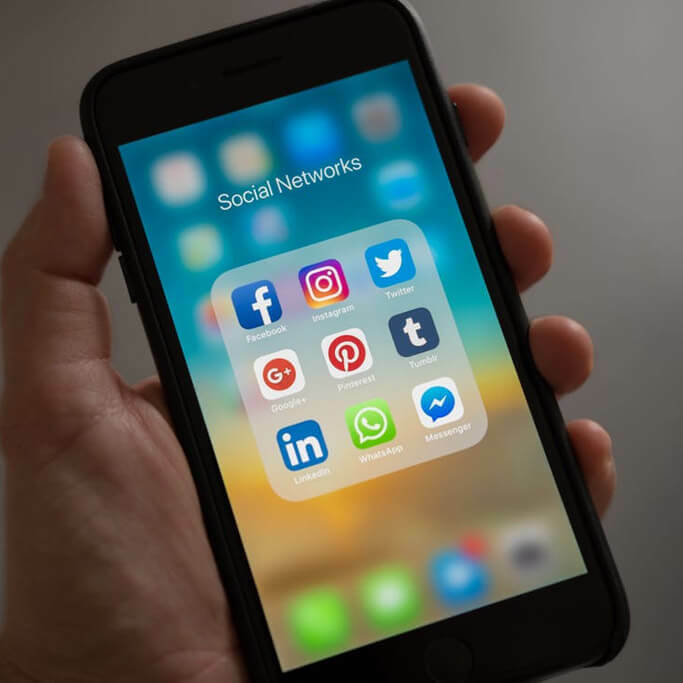 Hand holding a smartphone showing a Social Networks folder with Facebook, Instagram, Twitter, WhatsApp and other app icons.