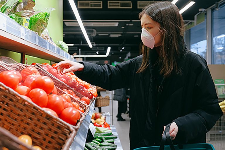 Woman wearing a face mask selecting tomatoes in a supermarket produce aisle.