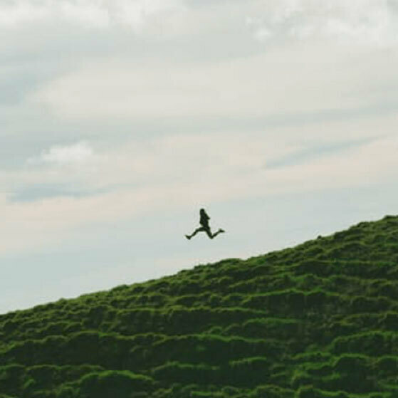 Person jumping above a grassy hill under a cloudy sky.
