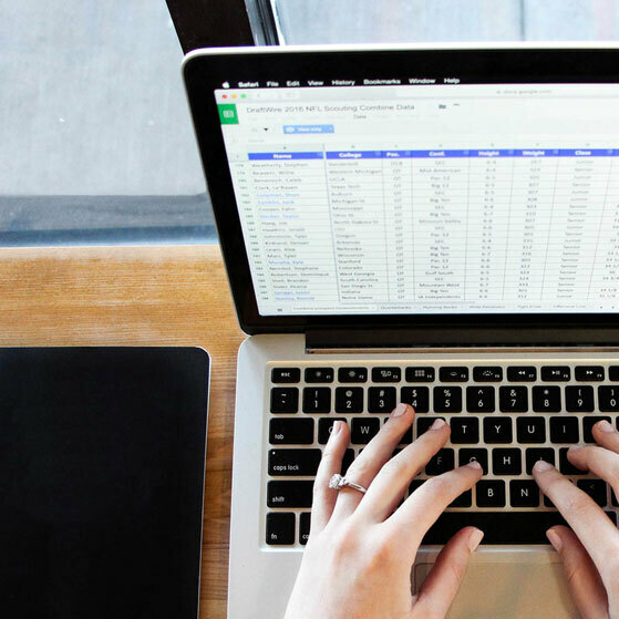 Hands typing on a laptop displaying a scouting data spreadsheet beside a tablet.