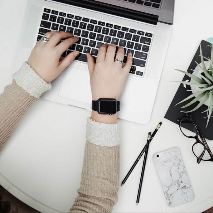Hands typing on a laptop at a white desk beside glasses, pencils, a phone, and a small plant.