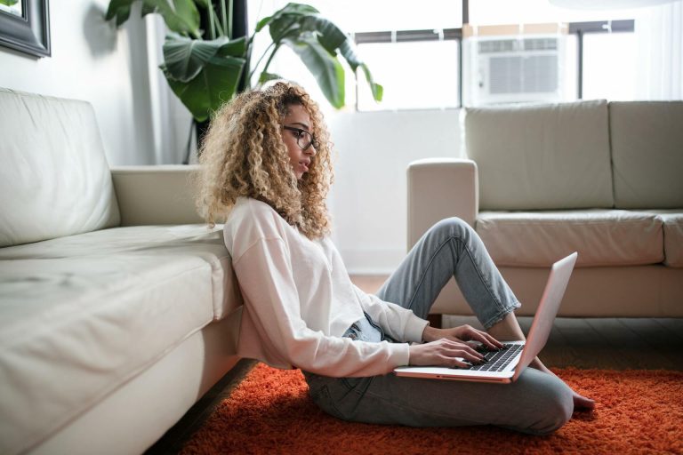 Woman working on a laptop while sitting on the floor beside a sofa.