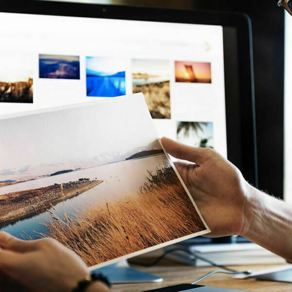 Hands holding a landscape print in front of a monitor with photo thumbnails.