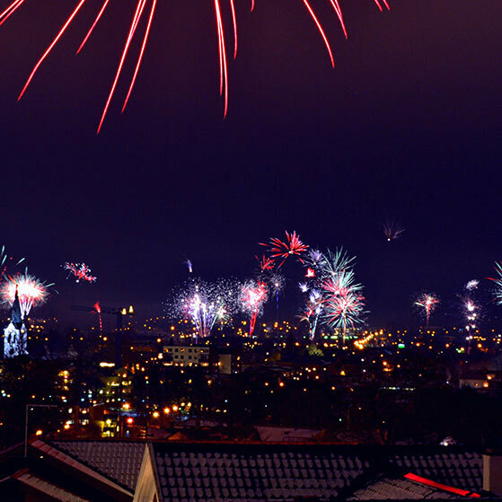Fireworks bursting over a city at night, with red streaks overhead and lights below.