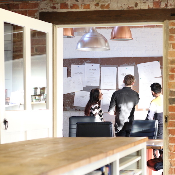 Three people discussing papers pinned to a notice board in a bright office meeting room.