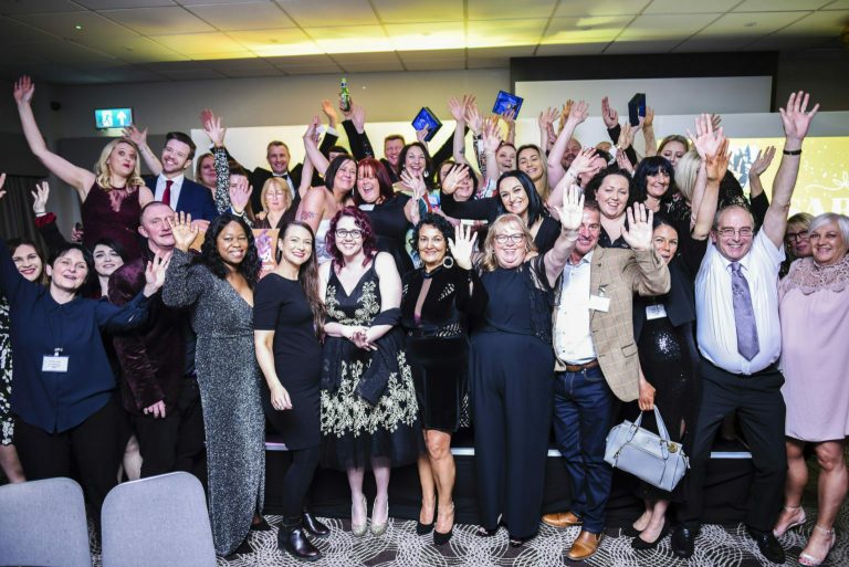 Conference attendees pose and wave on stage, holding awards and trophies.