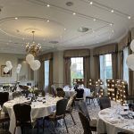 Elegant banquet room set for a celebration, with round tables, white balloons, and illuminated marquee letters.