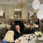 Guests seated at round tables with white balloons in an elegant dining room, with a harpist in the background.