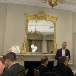 Man in a navy suit reading from a paper beside a fireplace at a formal dinner, with guests seated in front of a gilt mirror.