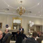 Man in a blue blazer speaks at a formal dinner as guests sit at tables with white balloons marked “W.”
