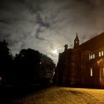 Moonlit church exterior with glowing windows and a grassy slope under cloudy night sky.