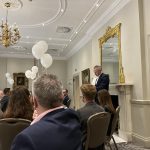 Guests seated in an elegant room as a man in a suit speaks beside the fireplace, with white balloons overhead.