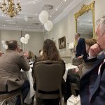 Guests seated at round tables listening to a speaker in an ornate banquet room with white balloons.