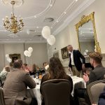 Guests seated at round tables applaud a speaker in a decorated banquet room with balloons and chandeliers.