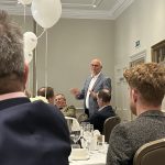 Man in a blue blazer speaks at a banquet table while guests listen in a formal dining room with white balloons.