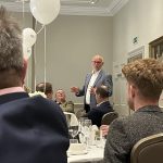 Man speaking at a formal banquet while guests listen at round tables with white balloons overhead.