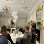 Guests seated at round banquet tables in an ornate dining room while a man speaks beside the fireplace.