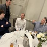 Four men chat around a round banquet table set with drinks and flowers in a formal room.