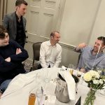 Four men chat around a round banquet table with drinks, flowers, and a place card reading Emily Kelsall.