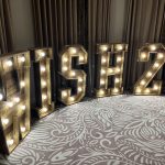 Illuminated wooden letters spelling WISH 2026 against curtains and a patterned carpet.