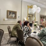 Guests seated at a round table chatting during a formal reception with white balloons and flowers.