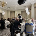 Guests seated at round tables in a banquet room while a waiter serves drinks with white balloons nearby.