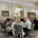 Guests seated at round tables at a formal dinner in a hotel-style room with white balloons and a gold mirror.