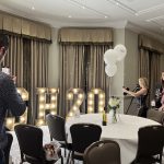 Guests at a round table pose beside illuminated H20 marquee letters and white balloons.