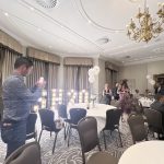 Guests mingle in an elegant banquet room with round tables, chandeliers, and illuminated marquee letters.