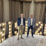 Two men stand in front of lit marquee letters reading “WISH20”.