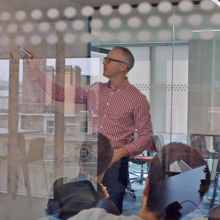 Man in a red check shirt writes on a glass whiteboard during a meeting.
