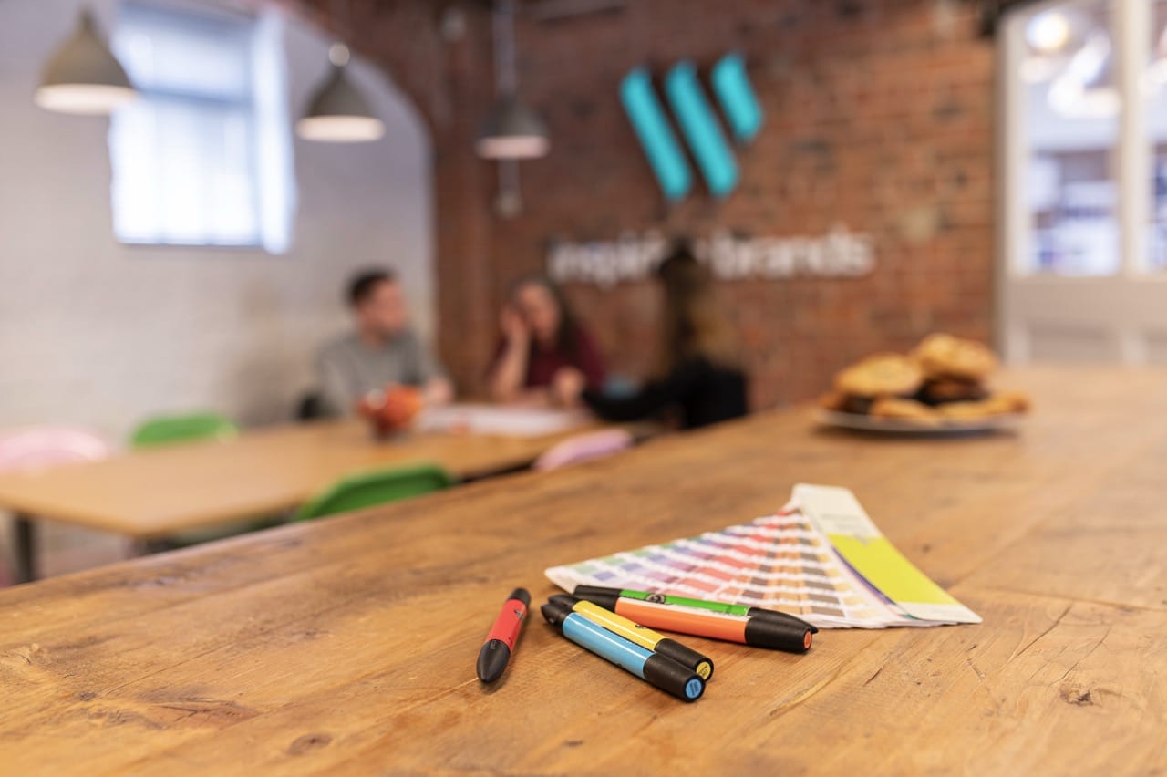 Markers and a color swatch fan on a wooden table in a creative studio, with people blurred in the background.